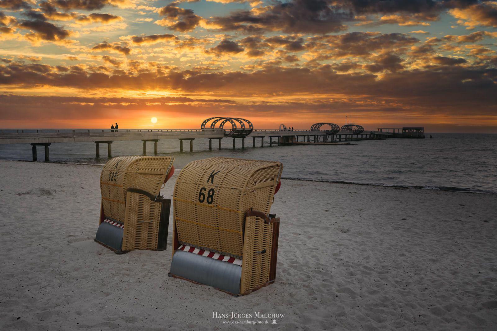 Strandkörbe am Strand bei Sonnenuntergang mit Blick auf eine Seebrücke.
