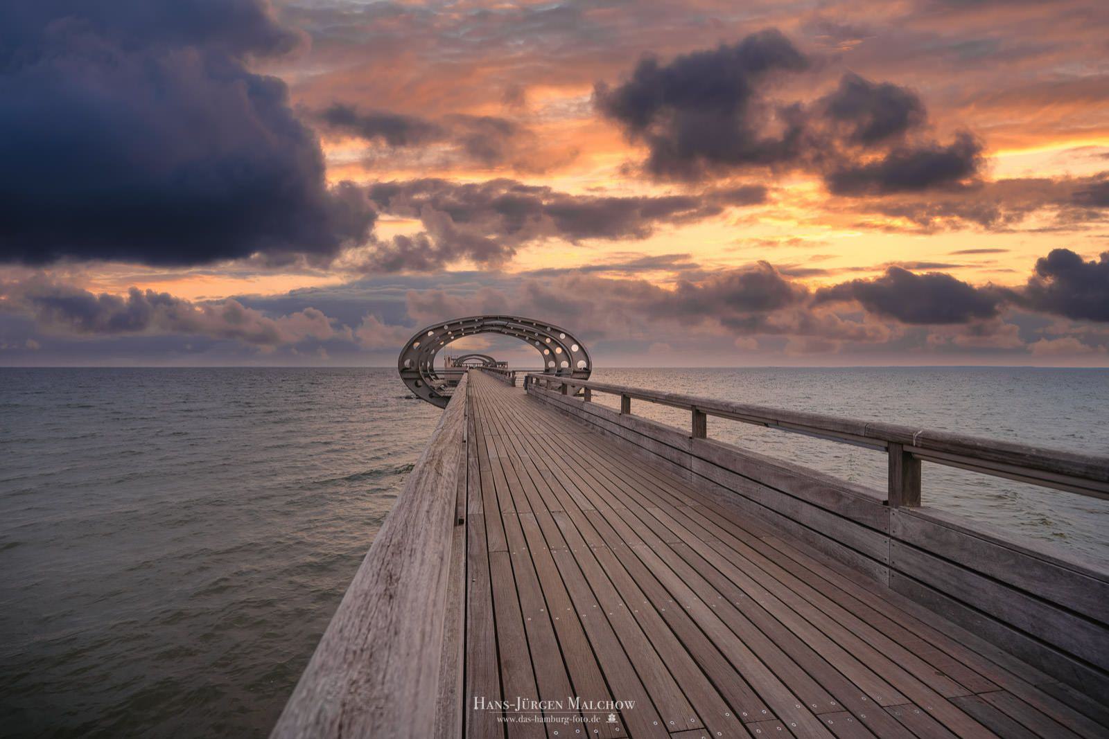Eine Holzpromenade führt auf dem Meer zu einer futuristisch anmutenden Metallkonstruktion unter einem dramatischen Sonnenuntergangshimmel.