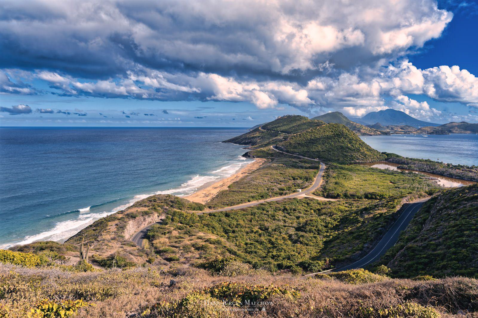 Eine kurvenreiche Straße schlängelt sich durch grüne Hügel an einer Küste mit einem Strand und dem blauen Meer.