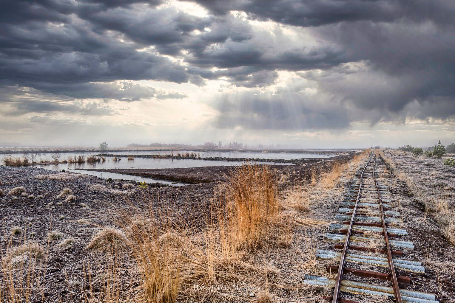 Verlassene Eisenbahnschienen, die sich durch eine karge, frostbedeckte Landschaft schlängern, unter einem dramatischen Himmel mit durchbrechenden Sonn