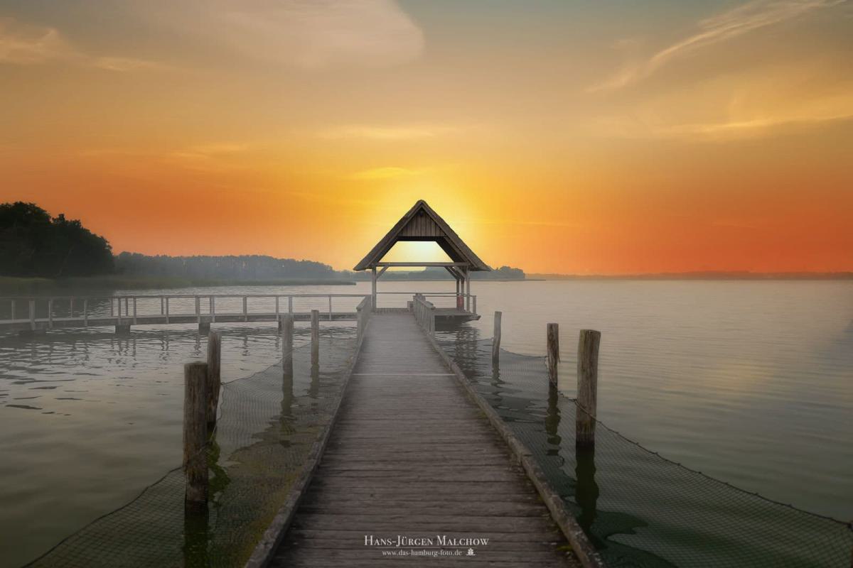 Holzsteg zu einer kleinen überdachten Anlegestelle am See bei Sonnenuntergang